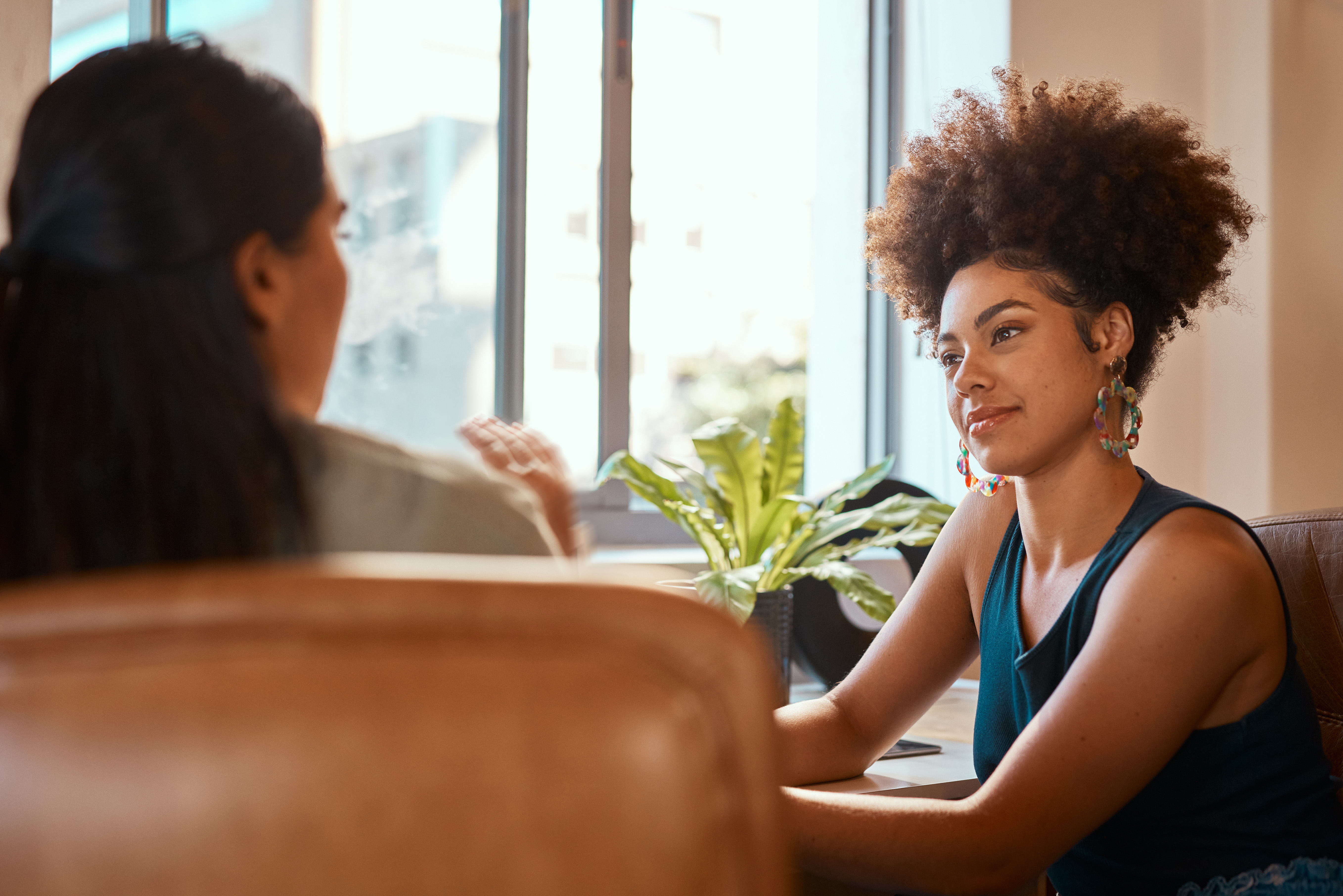 Black woman wearing large earings and talking to long hair woman sitting in brown leather office chair 