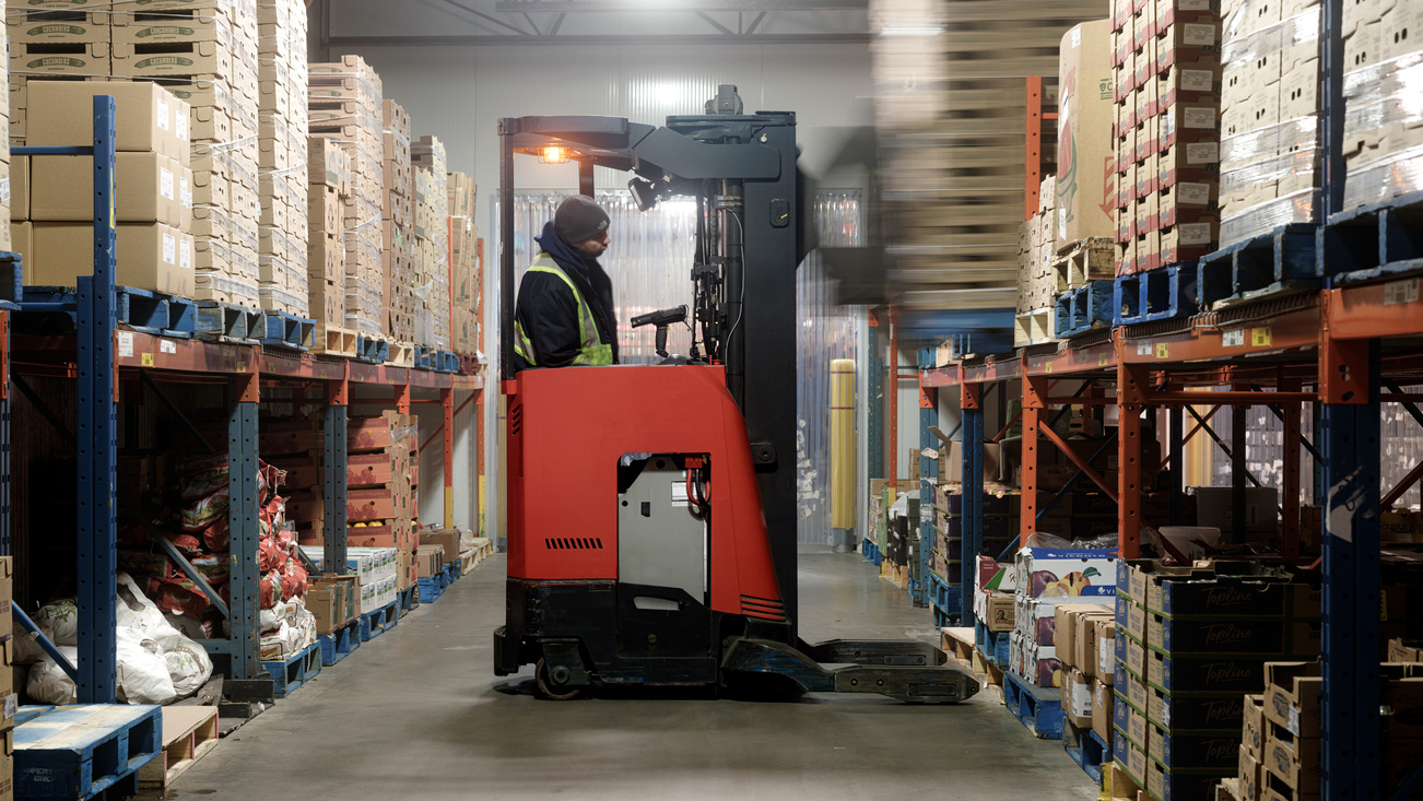 Warehouseman wearing fluorescent yellow safety jacket and driving a red forklift in a warehouse.