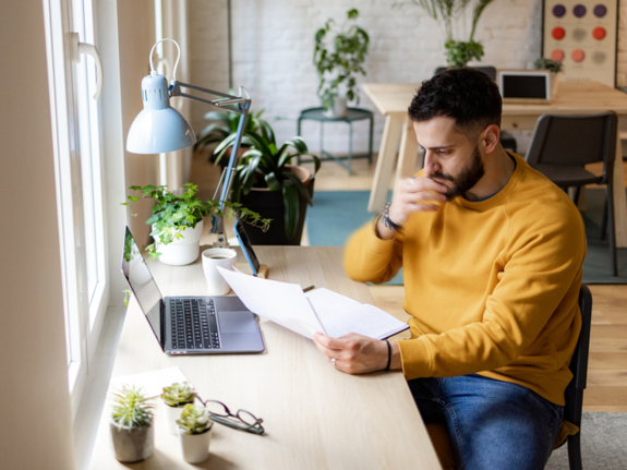 High angle side view of a handsome and successful young Caucasian professional male freelancer casually working from the comfort of his home, using a laptop and concentrating on his tasks